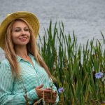 Happy woman with long blonde hair and straw hat smiling near a lakeside full of wildflowers.