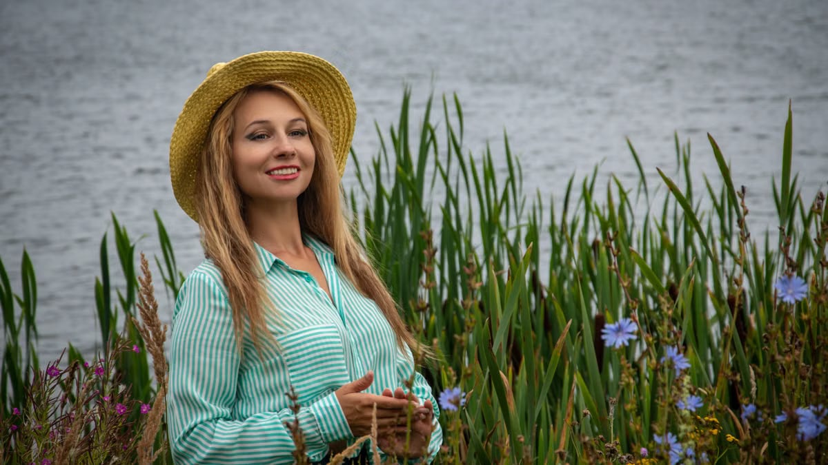 Happy woman with long blonde hair and straw hat smiling near a lakeside full of wildflowers.